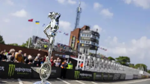 ISLE OF MAN TT A silver TT trophy in front of the TT grandstand, with pitlane and spectators in the background