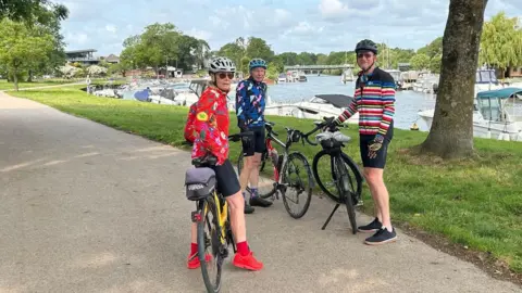 Lin Osborn saddling her bike and smiling at the camera. She is wearing a grey helmet, brown sunglasses, black cycling shorts and a red flowery cycling jacket. She is standing with two male friends who are also cycling and they have stopped by the River Stour in Christchurch