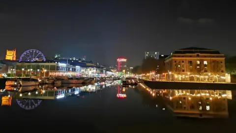A nighttime image of Bristol harbour - with a big expanse of water, which is in dark, and lit up buildings in the background