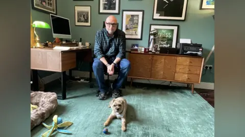 A man is sitting on a chair in an office, infront of him a small dog lies on the carpet with a ball