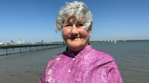 A woman with short grey hair wearing a pink shirt stands next to the water on a sunny day. You can see a city on the far horizon and there is a pier jutting out into the sea. 