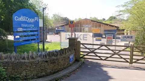 Google The exterior entrance to Bath Cats and Dogs Home in Somerset. There is a large blue sign above a curved brick wall which leads to a wide wooden gate. Behind it there is some metal fencing set up amid construction to convert an existing building into purpose built accommodation for animals.