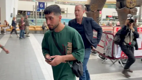 A young bearded man has turned away from journalist Andrew Harding and is heading to the Birmingham New Street station exit. The legs of the mechanical bull scuplture can be seen in the background and also in shot is a BBC cameraperson. 