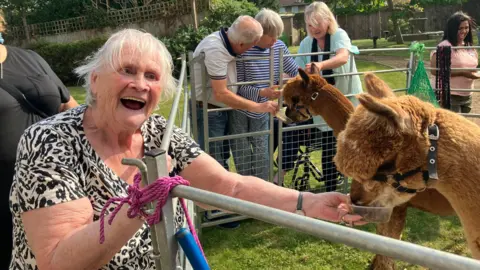 Shariqua Ahmed / BBC Valerie wearing a black and white T-shirt laughing while feeding a brown alpaca