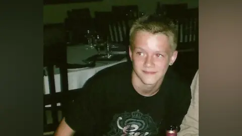 Perry Power A teenage boy standing up from a table. He is wearing a black t-shirt with a print on the front.