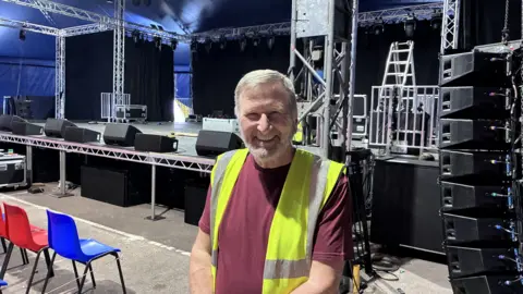 Ian Archer smiling and stood inside The Amp theatre. He is wearing a yellow high viz jacket and a dark red t-shirt underneath. He has white hair and a white beard. Behind him is a partially built stage with plastic seats placed in front of it.