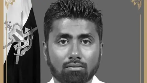 Maldivian diver Mohamed Mahdhee, with dark hair and beard, looks towards the camera. He is wearing a white shirt with epaulettes, medals and a nametag. There is a flag behind him.