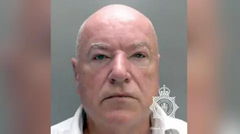 A police mugshot of Neil Foden, looking a the camera, and wearing a white shirt. He is balding, with grey hair. The mugshot has the North Wales Police logo on it.