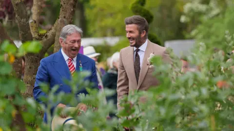 PA Media A man with short blonde hair and a light brown jacket and brown tie seen through green bushes laughing with a man with grey hair and wearing a blue jacket and red tie