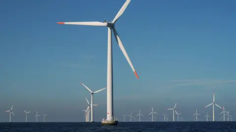 An array of about 30 offshore wind turbines. They are white with red-tipped blades. They are set against a blue sky.