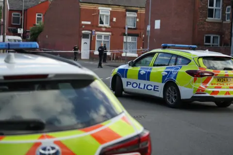 Image shows the scene in Manchester. Two police cars are in front of the image with a police cordon in the background. Three police officers can be see in the distance close to the cordon.