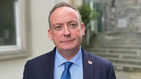 Deputy Lyndon Trott looking towards the camera, he has brown hair with flecks of grey and is wearing a blue suit with a Guernsey pin badge, with a blue shirt and metallic blue tie. 