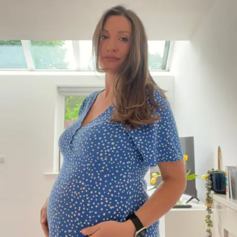 BBC/Linzi Kinghorn A pregnant woman standing up in a room with white walls and a window behind her. She has long light brown hair and is wearing a blue dress and white dots.
