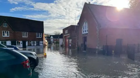 A street is completely flooded, with cars partially submerged and no pavements visible. There are red brick buildings, including homes.
