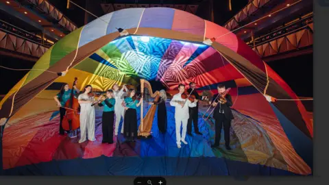 About eight musicians with different orchestral instruments, including a harp and a double bass, are standing inside a partially-inflated multi-coloured hot air balloon in a large auditorium. 