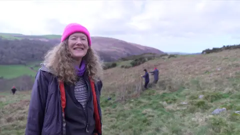 Woman with long, light brown, curly hair is standing on a hillside. She's wearing a bright pink beanie hat and a dark coloured coat. She's smiling at the camera. In the background you see three people on the hillside. Two are working on creating a dam from willow stakes. 