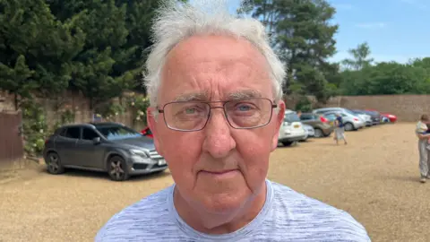 John Devine/BBC Ken Simpson is standing in a car park which has several cars parked in it against a wall. Simpson has short white hair and is wearing black framed glasses and a blue and white T-shirt.