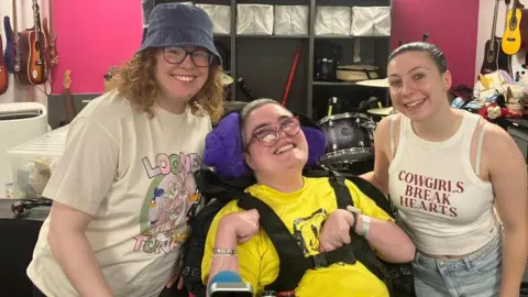 Three young women are in a band practice room. Guitars are on the walls behind them, a drum kit in the background. The woman in the centre of the pic is in a wheelchair. Each of the women is smiling.