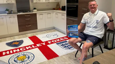 Darren Thompson Darren Thompson smiling with his hand in a fist and holding an atlas. He is sitting on a chair next to an England flag, which has four badges in each quarter and has Hinckley Foxes written in the middle.
