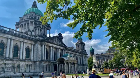 Kelly McCaughan Belfast city hall on a sunny day. The building is grey and there are lots of people walking around the gardens of it.