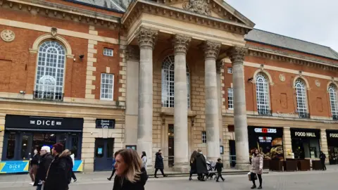 LDRS A grand building flanked by cafes and takeaways. The town hall has four pillars and large arched windows along the first storey. Shoppers are walking in front of the building.