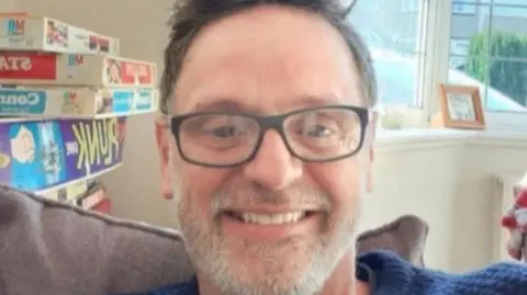 Chris Durham Chris Durham, a man with a shot grey beard , short brown hair and glasses smiles at the camera as he sits on a sofa. Behind him there is a window and on a shelf to his right there are a number of stacked boardgames