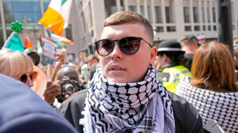 A close-up of a man in the midst of a crowd. He is wearing sunglasses and a black and white keffiyeh headdress around his neck like a scarf. A crowd is around him. You can see Irish tricolours in the background