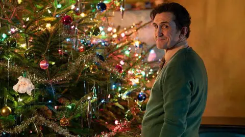 BBC/Neal Street Productions Stephen McGann sebagai Dr Turner, berdiri di dekat pohon Natal yang didekorasi dengan cerah dengan jumper hijau