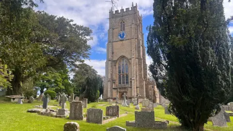 A church tower with a large clock in the top half looms above a graveyard, with old headstones dotted around the grass. A tall dark green tree stands to the front right of the church.
