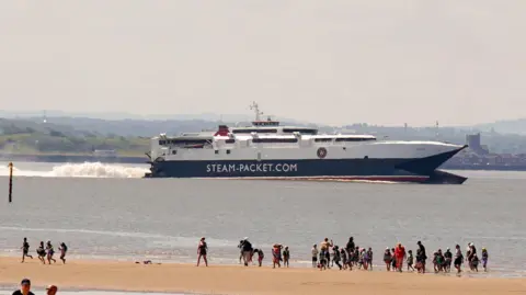 A ferry jets off while in the foreground people are stood on the beach watching it go by. 