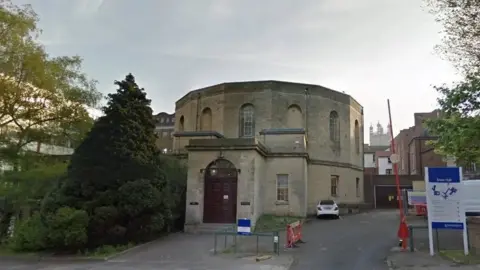 Google maps street view of Gloucester Crown Court. It is a beige, round building with a black door at the entrance. A blue and white sign is in front of the building showing a map. A white car can be seen parked in front of the court.