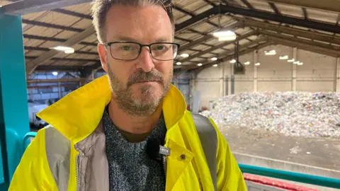 BBC Councillor Chris Watts, cabinet member for the environment and transport stands in front of a mountain of waste at a recycling plant. 