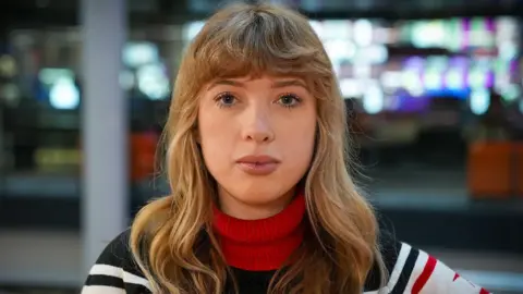 BBC Katie is sitting in chair in an office. She is looking at the camera. She has blonde hair and blue eyes. She is wearing a red, black and white jumper.