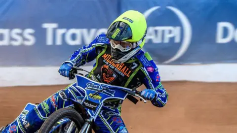 Getty Images A speedway racer steers his bike around a corner on a mud track. He wears a bright green helmet and blue racing suit with various sponsors written on it.