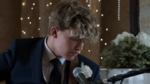 Fulwell Productions A screenshot of the Gavin & Stacey finale of Oscar Hartland, who plays Neil the baby, is wearing a suit and is singing into a microphone.