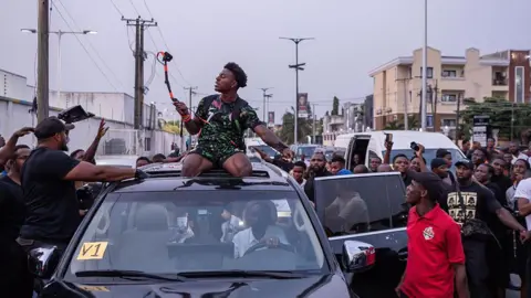 AFP via Getty Images IShowSpeed, sat atop a car, films himself and the crowd surrounding the vehicle. He wears a Nigerian football kit.