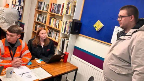 A man and a woman in Cadent branded uniforms sit behind a table. A member of the public stands in front of them.