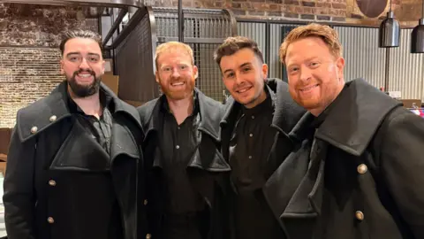 Boyd Rogers Four men dressed in black shirts and black coats with a large collars and large silver buttons. They are smiling. Pictured in front of a brick wall with metal sheet panelling. 
