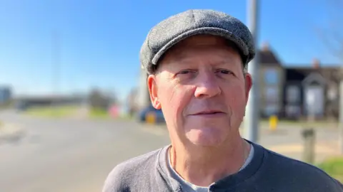 Lewis Adams/BBC Steve Thomas smiling on a sunny day. He is wearing a grey top and flat cap.