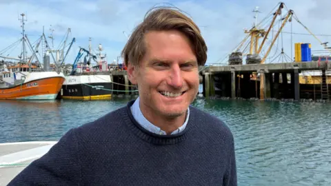 Chris Ranford smiling at the camera on a boat. Behind him is other vessels and the water. He is wearing a blue shirt with a knitted blue jumper over the top. 