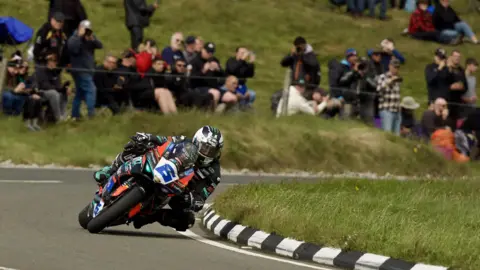 ISLE OF MAN TT Michael Dunlop on a largely black Supersport bike with the number six on the front. He's on the mountain section of the course with spectators watching from an embankment.
