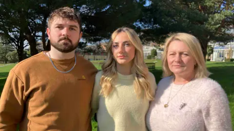 Fen Trudgian, wearing a tan jumper and a solid neck chain. He has a groomed beard and moustache and brown, lightly curly hair.