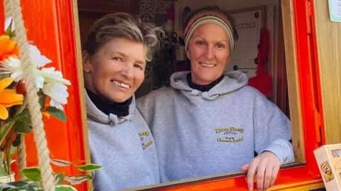 Nici Griffin Two women lean out of the serving hatch of a brightly painted narrowboat, smiling. Both wear grey hooded tops embroidered with the Three Sheep Cheese Boat name. Wheels of cheese and grapes are arranged on a wooden counter in front of them.