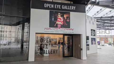 External view of the gallery, which has a large glass entrance that says "Open eye Gallery" in black text on the window. Above it is a poster advertising the exhibition