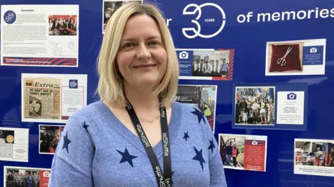 BBC/ Jo Burn A middle-aged woman with shoulder length blonde hair wears a blue jumper with stars on it and smiles at the camera in front of a board of archive photographs of the Channel Tunnel 