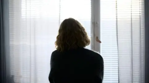 A person looking out of a window. We can see the person from the back. There are blinds and net curtains across the window