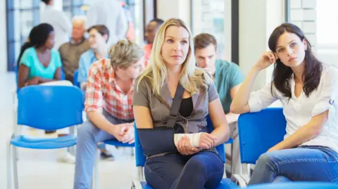 A full waiting room. One woman sits at the front with her arm bandaged and in a sling.