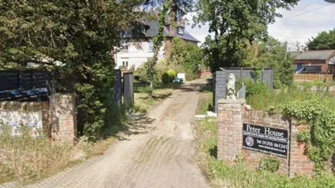 Exterior of a large white house surrounded by a brick wall on either side of the house. There are bushes and small statues at the front of the house with a large tree hanging over the front. There is a small black sign at the front with white writing saying Peter House and the telephone number.