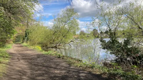 A view of Poynton Pool on a sunny spring day. The image shows the pool on the right, with a footpath on the left. There are a number of trees along the bank of the pool.