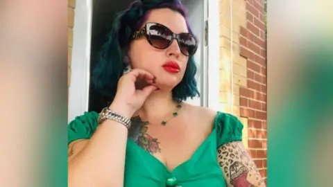 Tania Williams, is sitting outside and posing for the camera. She is wearing a green dress with sleeves that sit just across her shoulders. She is not looking at the camera and is instead looking off towards the right. Her arm is rested on the side of her face and she is wearing a pair of tortoise shell sunglasses and red lipstick.
